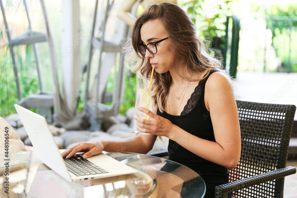 beautiful young girl in glasses sits with a laptop at a table in a summer cafe. young woman blogger drinks a cold cocktail in a cafe in the park. business woman looking through e-mail in a summer cafe beautiful young girl in glasses sits with a laptop at a table in a summer cafe. young woman blogger drinks a cold cocktail in a cafe in the park. business woman looking through e-mail in a summer cafe