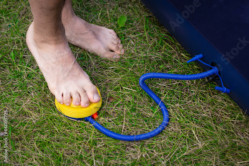 Male legs swing the inflatable mattress using a foot pump. A man stands barefoot on the grass and presses on the pump. photo