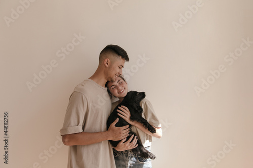 Young family with little black dog against light background. Cute young boy in beige loungewear hugging pretty lady holding puppy indoor photo