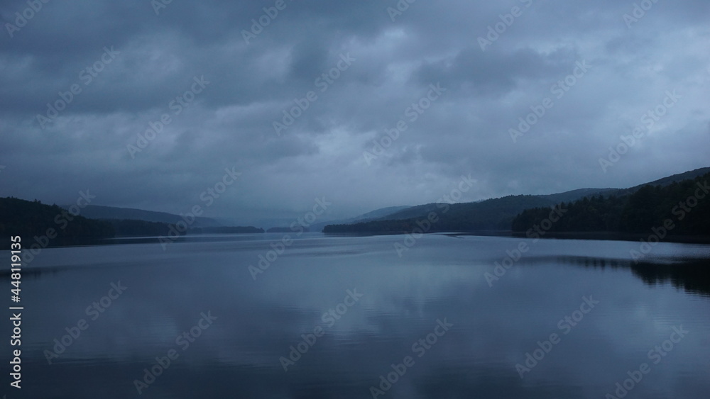Clouds hover over a still lake on a grey evening.  Mountain background.