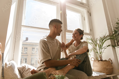 Lovestory portrait of young couple at home. Pretty pair of trendy people hugging each other, smiling and sitting near light window and decorations
