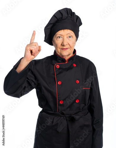 woman chef in black uniform. apron, shirt, hat. elderly woman. the lady looks at us, raised her finger up as a sign of warning, attention. white background, isolated