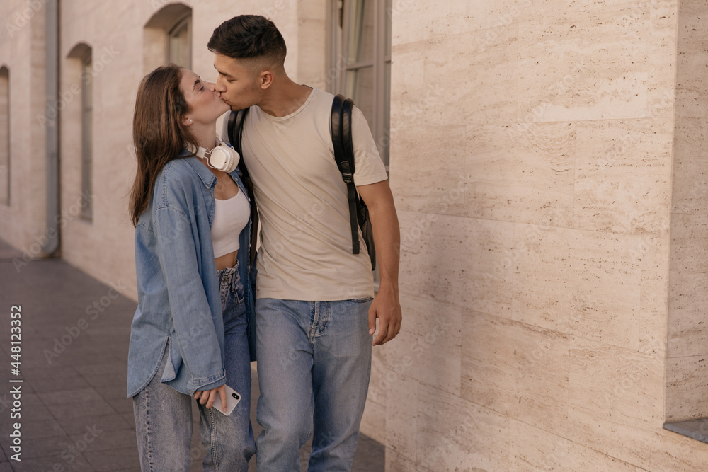 Love, relationship, happiness concept. Young dark-haired girl wearing denim shirt kissing boyfriend in beige t-shirt and jeans against light building background