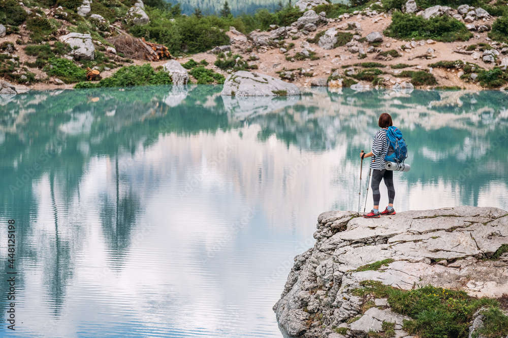Fototapeta premium Backpacker woman with backpack and trekking poles enjoying the turquoise Lago di Sorapiss 1925m altitude lake view during mountain walking in Dolomite Mountains, Italy. Active people in nature concept