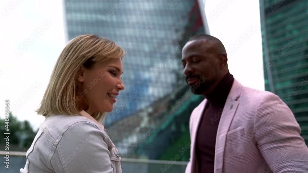 a bearded African-American man in a pink blazer and a blonde in a white jacket are dancing merrily against the background of high-rise buildings a bearded African-American man in a pink blazer and a blonde in a white jacket are dancing merrily against the background of high-rise buildings