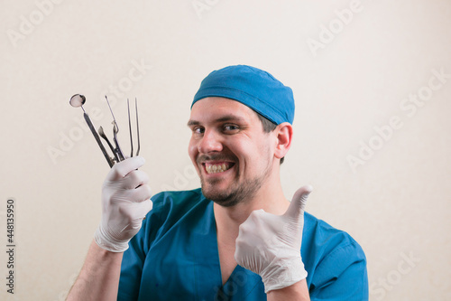Young smiling doctor with medical instruments in his hand and thumb up in blue surgeon suit isolated
