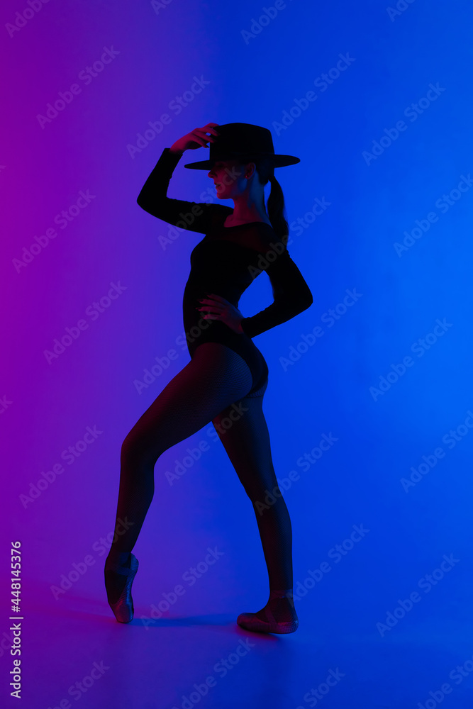 Contemporary ballet dancer woman in hat posing on multi-colored gradient pink blue background. Professional ballerina in sexy costume of femme fatale, solo performance.