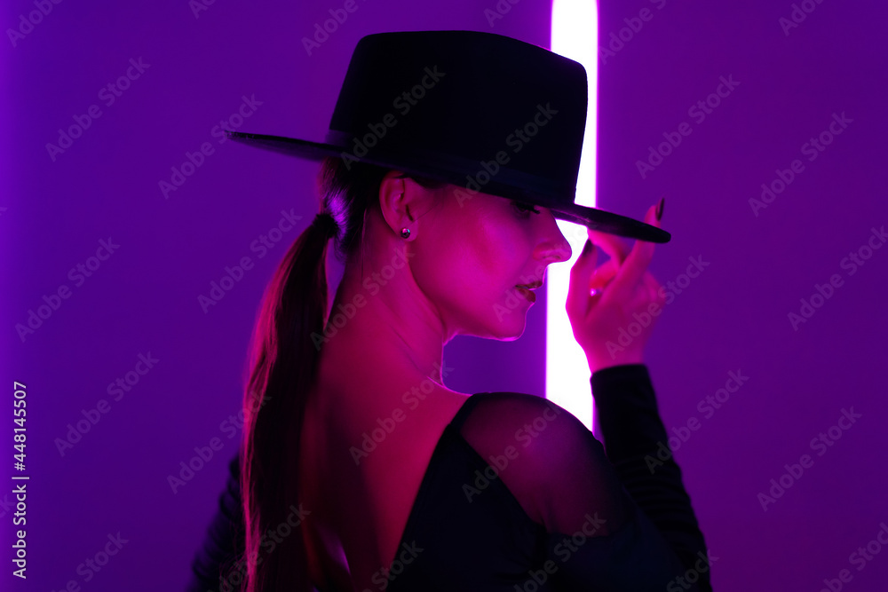 Alluring woman in hat between flashing colored neon lights, studio background. Femme fatale, stylish outfit of ballet dancer.