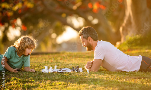 happy family of dad and son boy playing chess on green grass in park outdoor, concentration