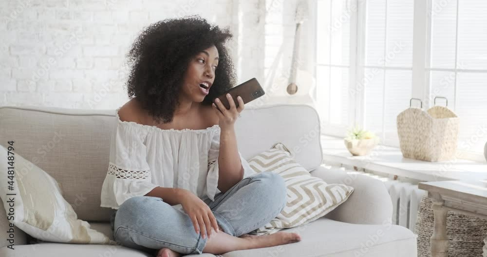 Young smiling biracial woman recording audio message to friend using smartphone sitting on grey sofa at home close view. Voice chat and clubhouse concept. Using digital assistance on mobile phone. Young smiling biracial woman recording audio message to friend using smartphone sitting on grey sofa at home close view. Voice chat and clubhouse concept. Using digital assistance on mobile phone.