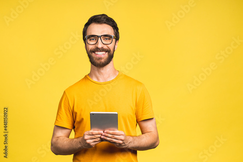 Happy young man in casual standing and using tablet computer isolated over yellow background.