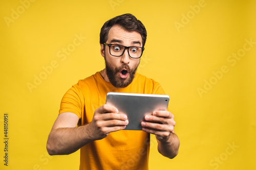 Amazed happy bearded man using digital tablet looking shocked about social media news, astonished man shopper consumer surprised excited by online win isolated over yellow background. photo