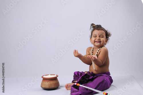 adorable Indian baby in krishna kanha or kanhaiya dress posing with his flute and dahi handi (pot with curd) on white background. sitting pose photo