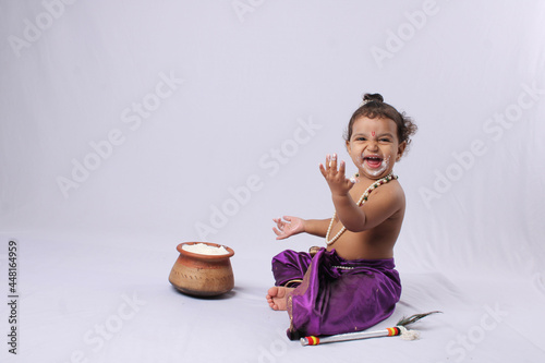 adorable Indian baby in krishna kanha or kanhaiya dress posing with his flute and dahi handi (pot with curd) on white background. sitting pose photo