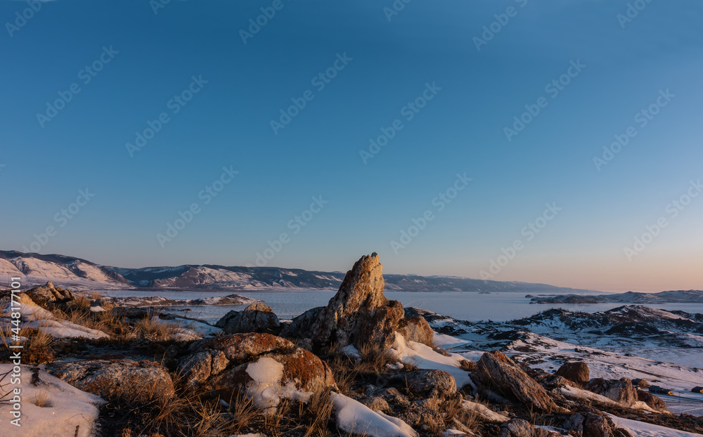 Early winter morning in Siberia. The frozen lake is surrounded by mountains. There are picturesque boulders with lichens on the shore. There is snow and dry grass around. The golden hour. Baikal