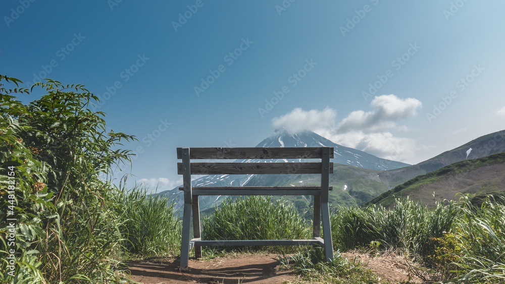 Observation deck with a view of the Vilyuchinsky volcano. There is a wooden bench among the grass. White clouds float over the snow-capped mountain top. Clear blue sky. A summer day. Kamchatka