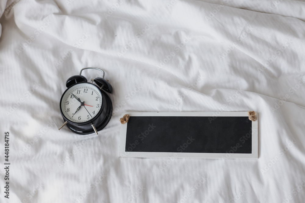 Black alarm clock showing 7 oclock on the bed with blank sign board for mockup Black alarm clock showing 7 oclock on the bed with blank sign board for mockup