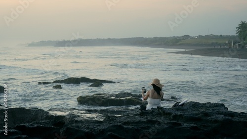 Anonymous woman with black hair and a straw hat sitting alone on the rocks of a reef while the ocean tide is waving around after sunset. Atmospheric twilight with a single female person enjoying beach