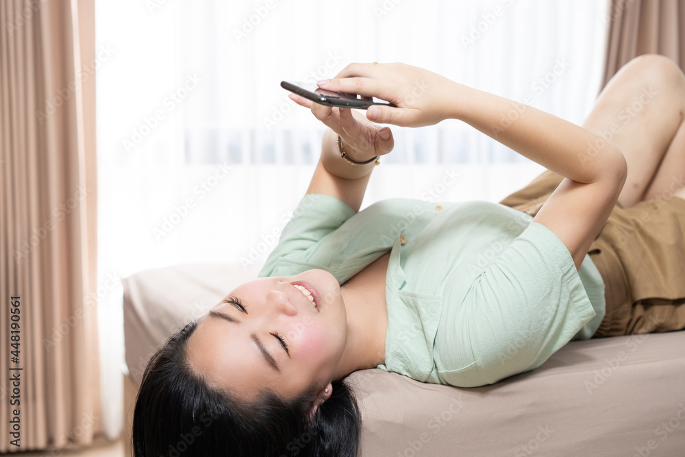 Beautiful Asian woman is lying down and playing mobile on the bed in her bedroom with a pastel green - brown color theme. Beautiful Asian woman is lying down and playing mobile on the bed in her bedroom with a pastel green - brown color theme.