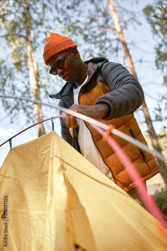 Black man setting tent on meadow in forest. Stay for sleep on open fresh air. Skilled for camping, hiking and travelling alone. Relaxation in nature, unique landscape, find peace. Adventure concept