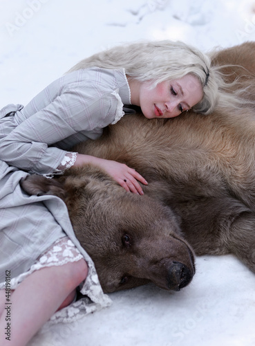Beautiful white haired girl in vintage dress hugging brown bear in winter forest