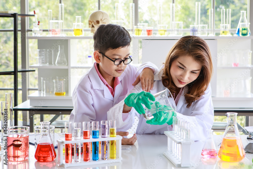 Attentive Asian schoolboy watching female tutor pouring liquid into flasks while conducting experiment with colorful reagents during chemistry lesson in laboratory