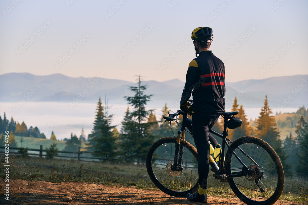 Back view of male cyclist in cycling suit standing with bike in the morning. Man bicyclist wearing safety helmet while enjoying bicycle ride in mountains. Concept of sport, biking and active leisure.