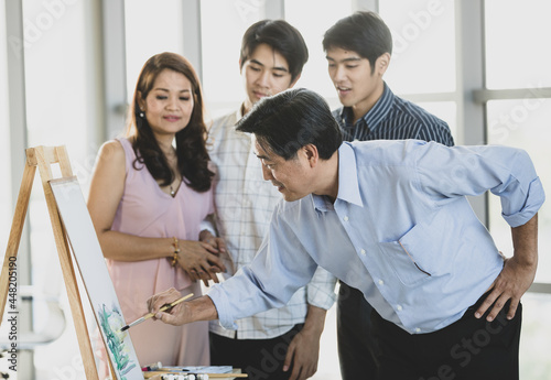 50s Asian father happily painting coloring on canvas and easel as hobby while two handsome sons and beautiful wife laughing with happiness and fun together during the weekend holidays at cozy home.