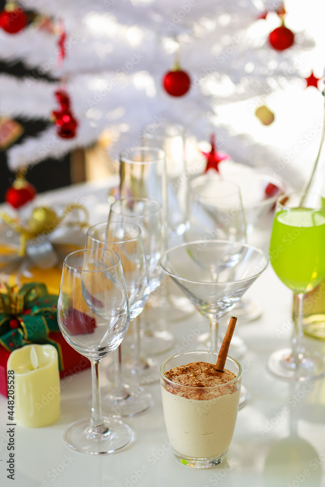 Tasty delicious milk cream eggnog drink with gingerbread in glass on table in front empty tall wine glasses present gift boxes white chocolate candles green apple punch and decorated Christmas tree Tasty delicious milk cream eggnog drink with gingerbread in glass on table in front empty tall wine glasses present gift boxes white chocolate candles green apple punch and decorated Christmas tree