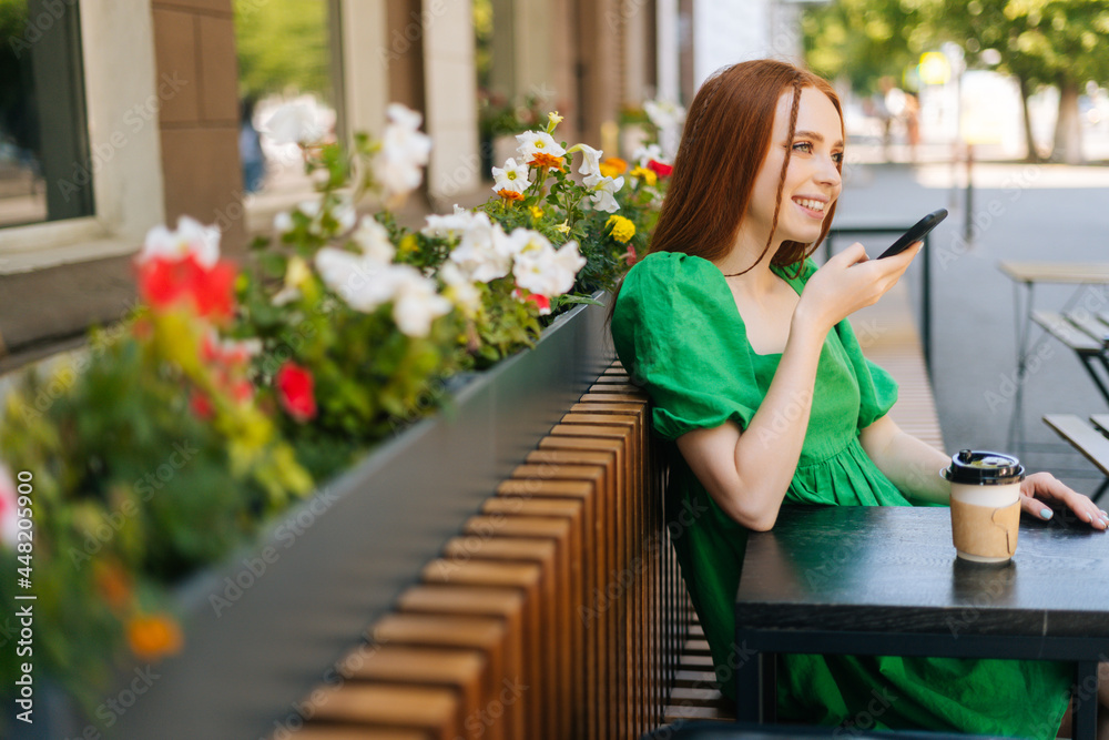 Side view of smiling redhead young woman recording audio voice message on cellphone sitting at table with coffee cup in outdoor cafe terrace in sunny summer day.