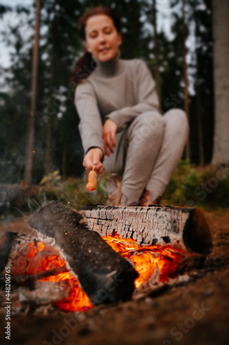 Young female tourist cooking barbecue sausages on a campfire. Selective focus on fire.