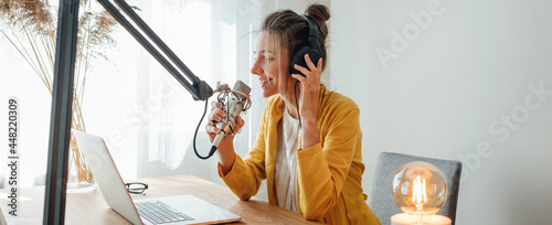 Cheerful woman podcaster recording her voice into microphone. Female radio host streaming podcast using microphone and laptop at his home studio photo