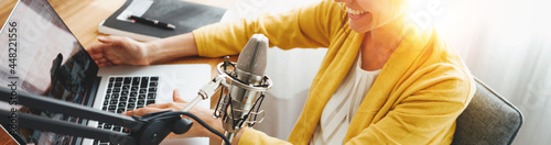 Cheerful woman podcaster recording her voice into microphone. Female radio host streaming podcast using microphone and laptop at his home studio. Wide image photo