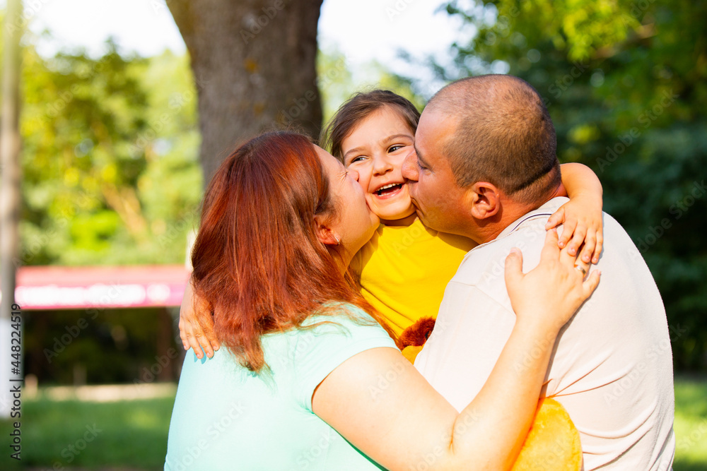 Lovely family are spending wonderful sunny day time together in the park holding the girl in their hands and kissing to each other.