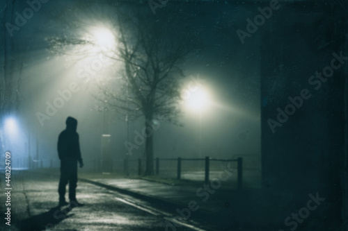 A spooky hooded man standing underneath a street light. On a foggy winters night in the city