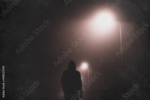 A hooded man standing underneath a street light, back to camera. On a foggy winters night. With a blurred, textured edit.