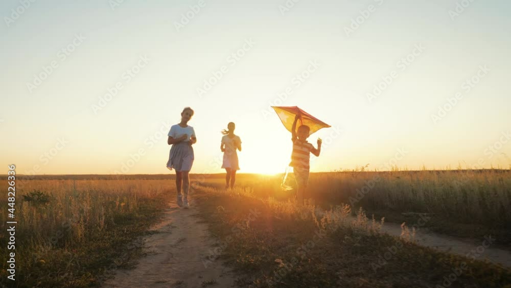 Happy young mother with a children play kites together on park. Boy running with flying kite in hand. Silhouette of happy people running at sunset in the park. Happy parents childhood dream concept.
