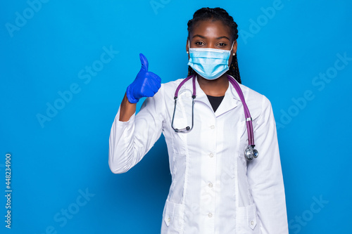 Excited african american doctor woman isolated on blue background in studio. Female doctor in white medical gown showing thumb up.