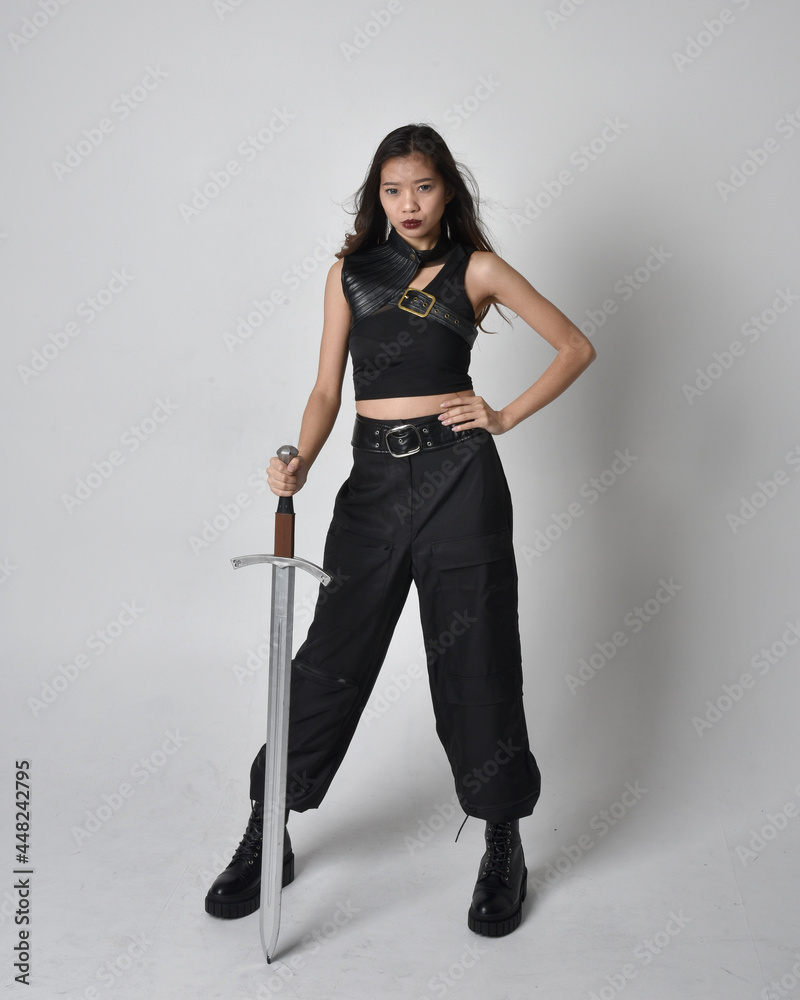 
Full length portrait of pretty young asian girl wearing black tank top, utilitarian  pants and leather boots. Standing pose  holding a sword, isolated against a  studio background.