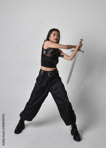  Full length portrait of pretty young asian girl wearing black tank top, utilitarian pants and leather boots. Standing pose holding a sword, isolated against a studio background.