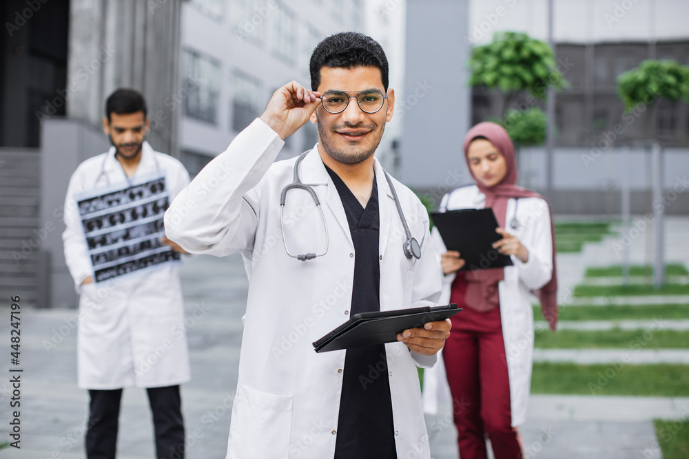 Coworking outdoors of hospital. Young handsome arab hindu man doctor in glasses using tablet device, posing to camera with smile. Multi ethnic male and female physicians working on the background Coworking outdoors of hospital. Young handsome arab hindu man doctor in glasses using tablet device, posing to camera with smile. Multi ethnic male and female physicians working on the background