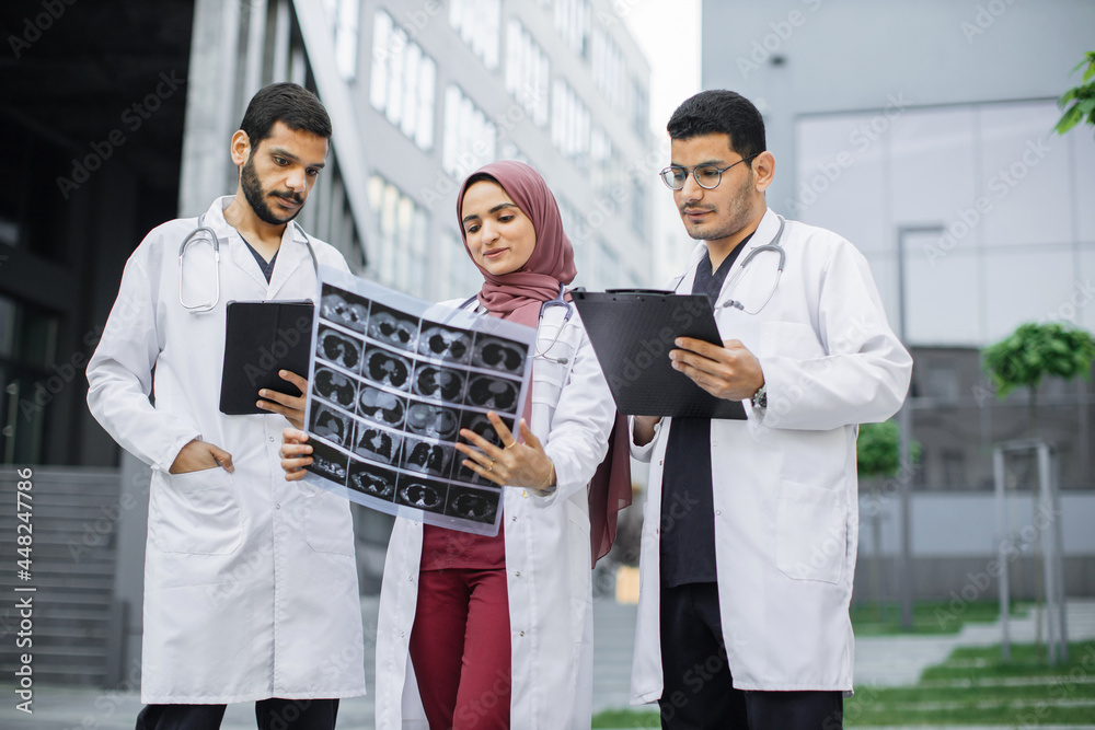 Team of professional three male and female Arabian doctors examining patient's tomography scans while standing outside the modern hospital building. Doctors discussing x-ray image Team of professional three male and female Arabian doctors examining patient's tomography scans while standing outside the modern hospital building. Doctors discussing x-ray image