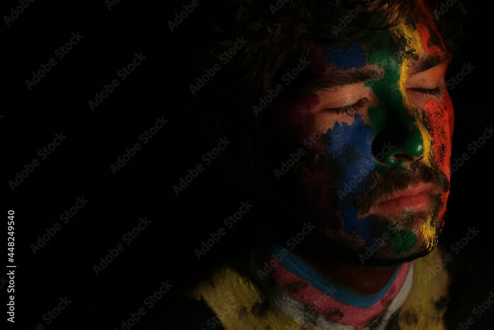 Portrait of young man face painted as lgbtq progress pride flag with direct lighting on face with dark black copy space empty background *4