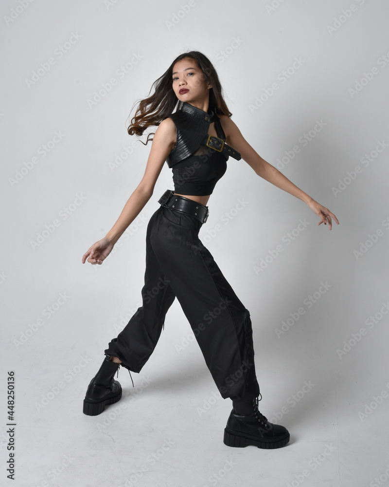 Full length portrait of pretty young asian girl wearing black tank top, utilitarian  pants and leather boots. Standing pose with gestural hands, isolated against a  studio background.