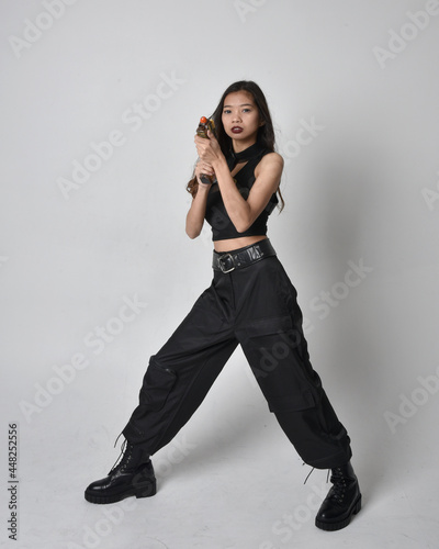 Full length portrait of pretty young asian girl wearing black tank top, utilitarian pants and leather boots. Standing pose holding a gun, isolated against a studio background.