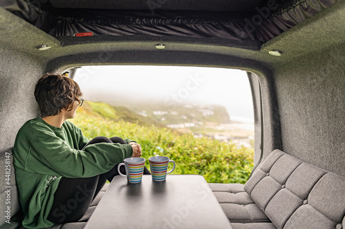 Photo of an attractive young female drinking coffee and sitting on a sofa inside her campervan while enjoying the views on a foggy day