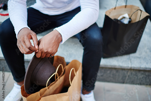 Selective focus on black male hands holding fashionable stylish hat near paper bags after buying purchase, cropped image of dark skinned man shopaholic resting on stairs during weekend shopping