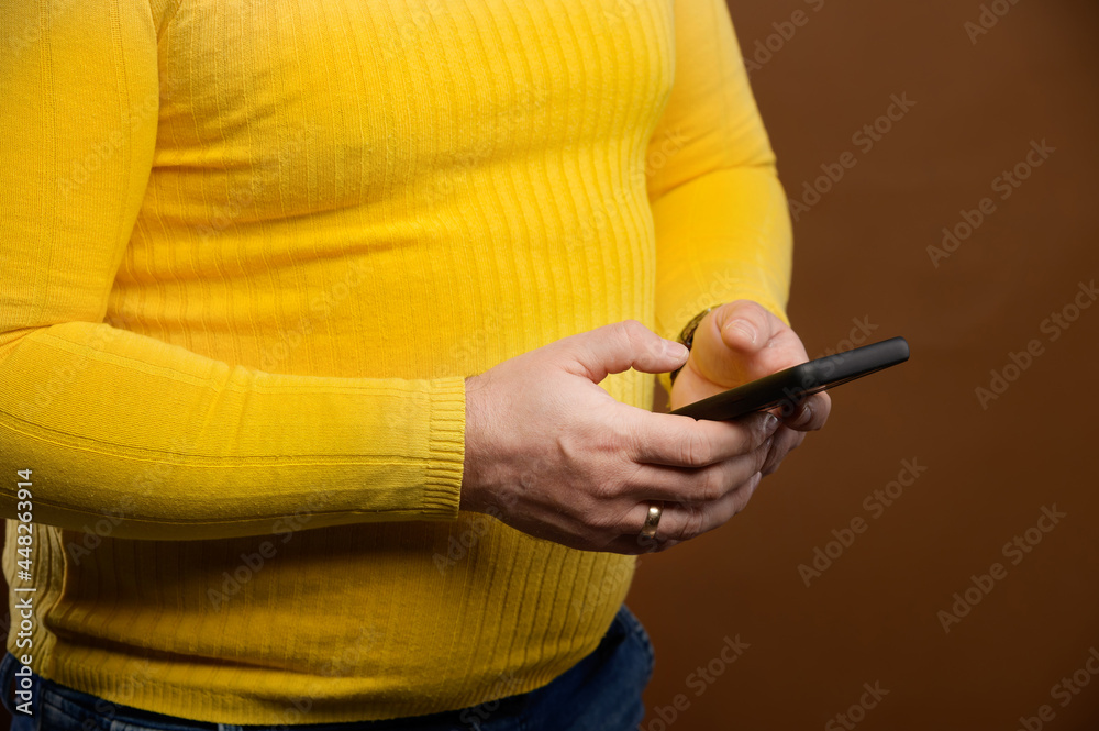 Close-up of a plump middle-aged man in a yellow casual jacket uses his phone while typing or surfing the Internet. Studio shot on brown background