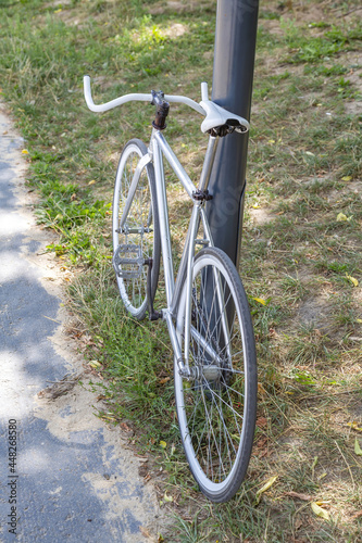 Bicycle with white wheels is parked by an iron pillar in a city summer park