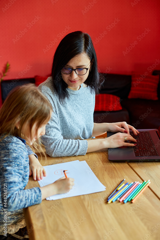 Fototapeta premium Mother working in her home office on a laptop, her daughter sits next to her and draw